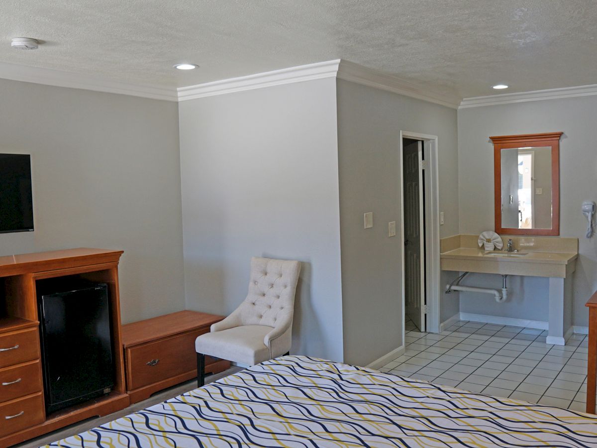 The image shows a tidy hotel room with a TV, dresser, chair, bed, sink area, and wall mirror. The room appears to have neutral colors.