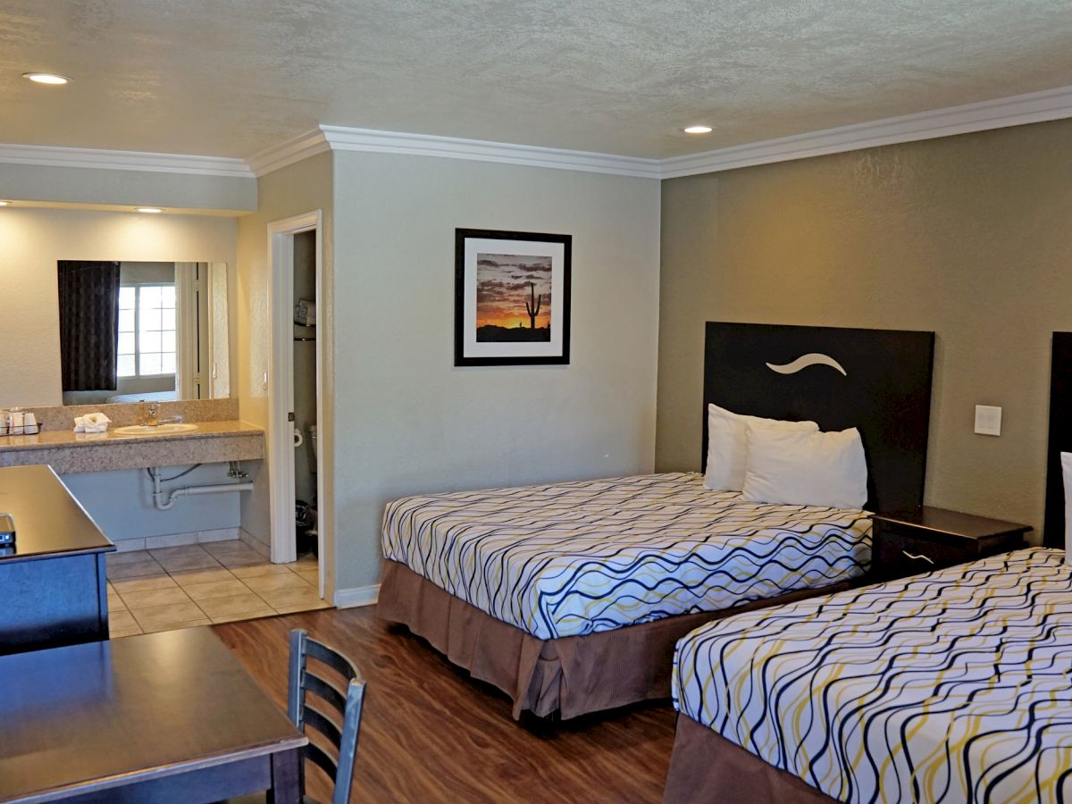 A hotel room with two beds, a desk, a TV, and a sink area leading to a bathroom. There is a picture on the wall above the beds.