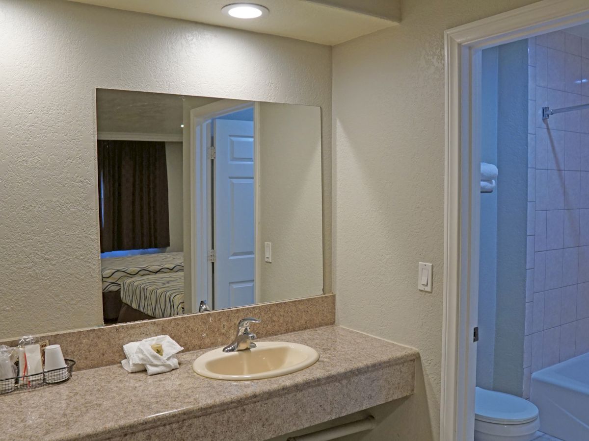 The image shows a bathroom with a large mirror, a sink, and various toiletries on the counter, adjacent to a toilet and a shower/tub with a curtain.