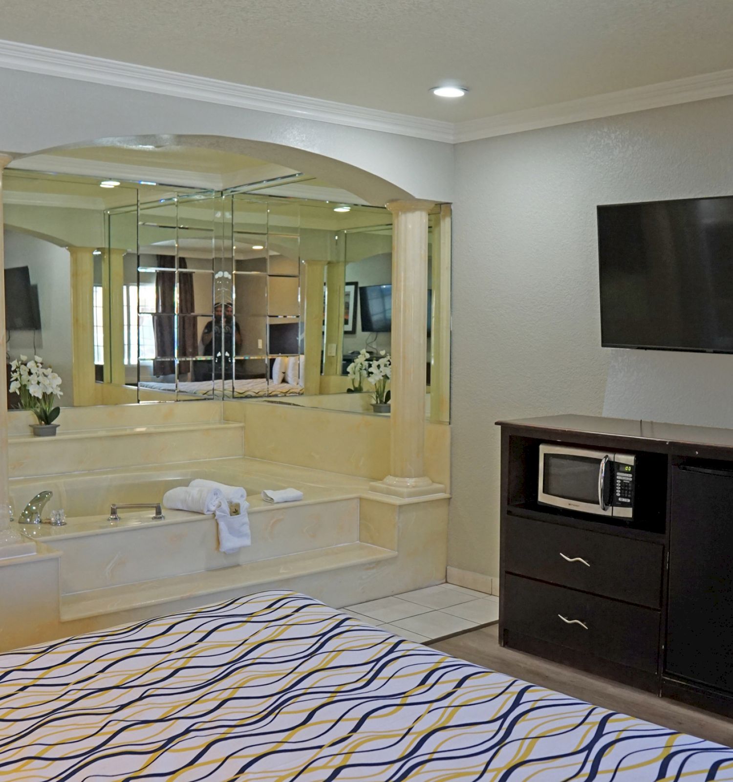 This image shows a hotel room with a bed, a wall-mounted TV, a jacuzzi tub with mirrors, a microwave, a coffee maker, a desk, and a bathroom.