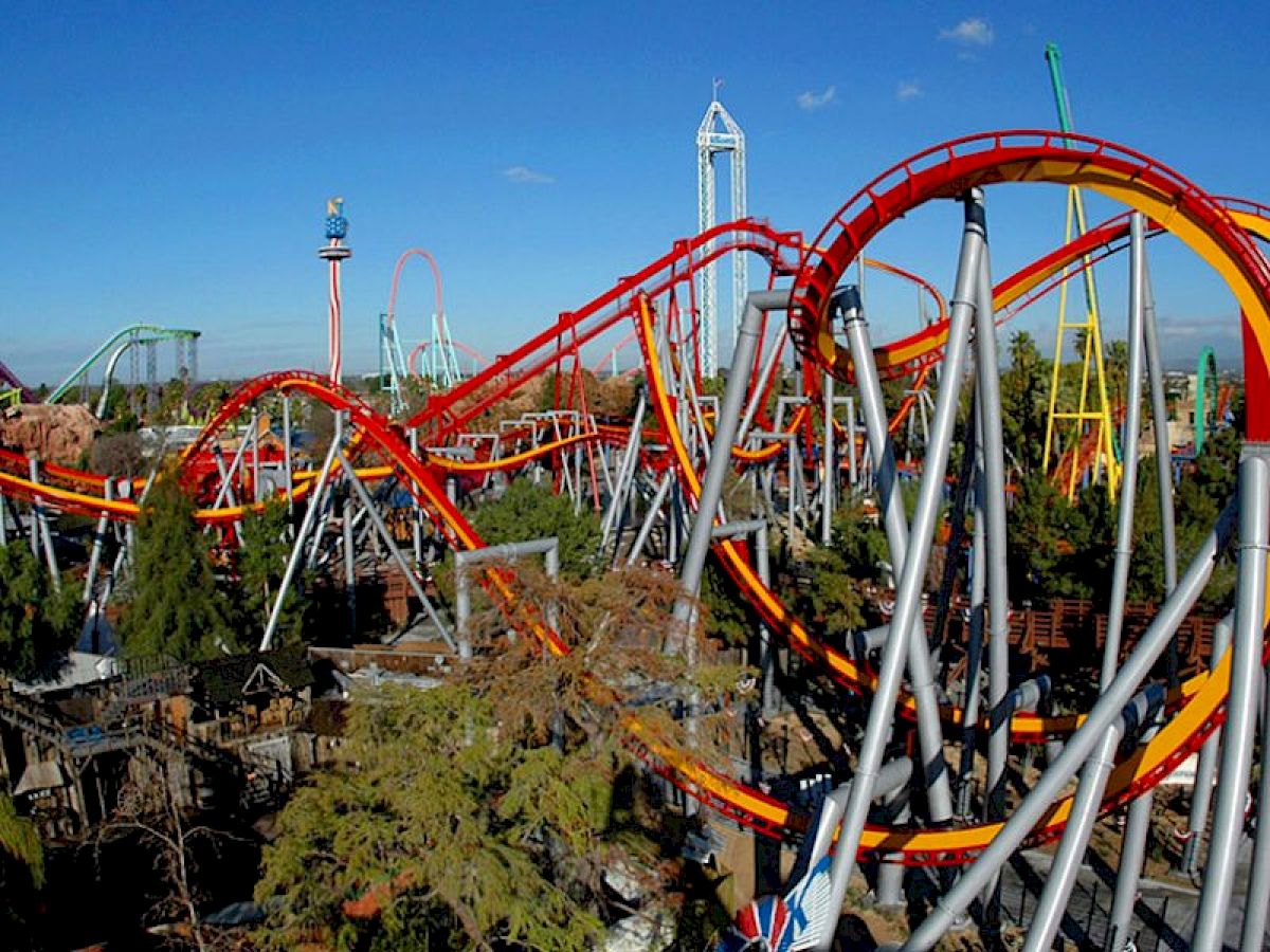 An image of a theme park with multiple roller coasters and rides with red and yellow tracks surrounded by trees and other attractions.