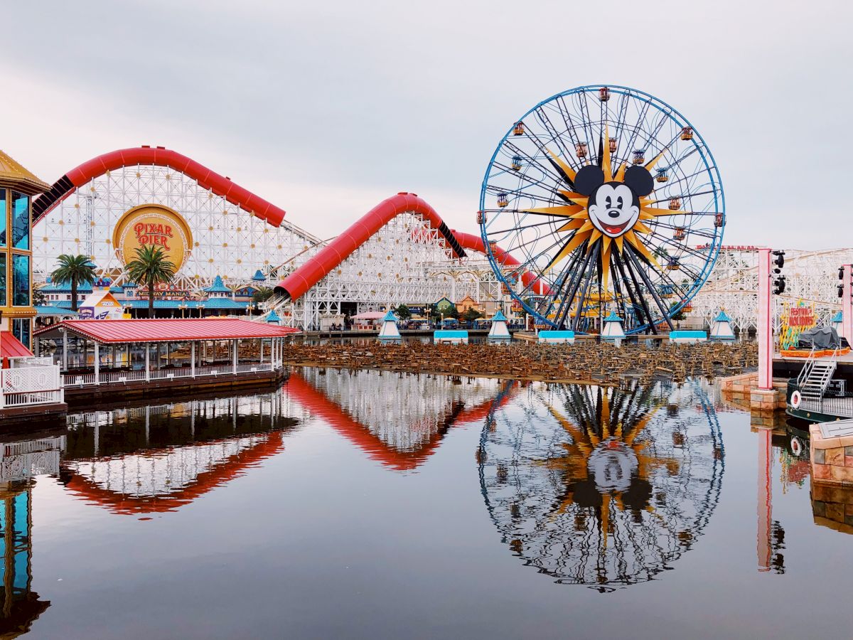 The image depicts an amusement park with a Mickey Mouse-themed Ferris wheel, a roller coaster, reflection in water, and various attractions visible.
