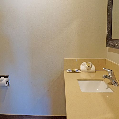 The image shows a bathroom with a toilet, a wall-mounted toilet paper holder, a countertop with a sink, a faucet, a mirror, and toiletries.