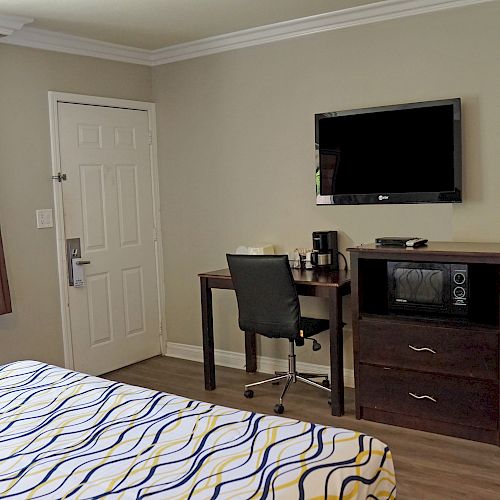 The image shows a hotel room with a bed, TV, desk, chair, mini-fridge, coffee maker, and a window with brown curtains next to a white door.