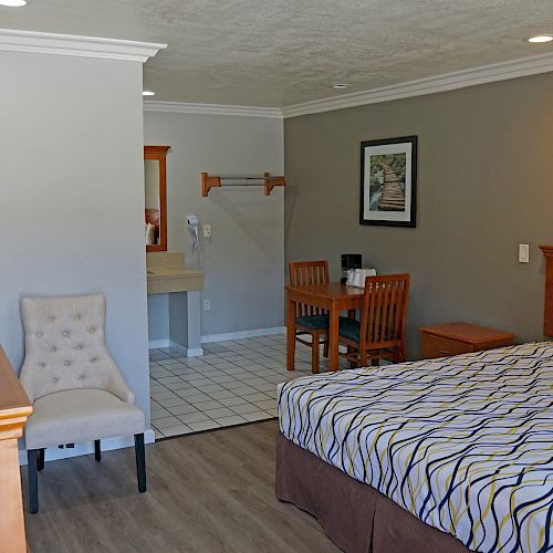 The image shows a hotel room with a bed, a small table and chairs, a chest of drawers, a mirror, a sink area, and simple wall decor.