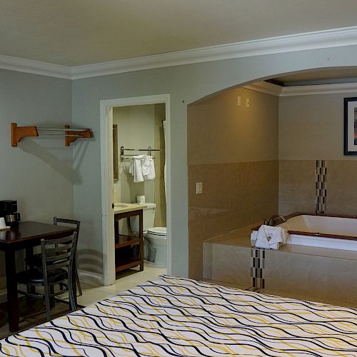 A hotel room features a bed, a TV mounted on the wall, a table with chairs, a dresser, an open bathroom, and a large bathtub in an alcove.