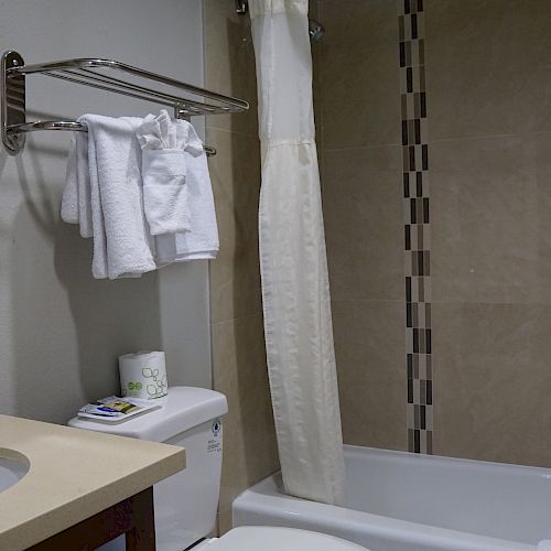 This image shows a clean, modern bathroom with a sink, mirror, toilet, towels on a rack, and a bathtub with a shower curtain.
