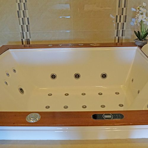 This image shows a luxurious whirlpool bathtub with wooden trim, surrounded by beige tiles and adorned with a potted plant and rolled towels.