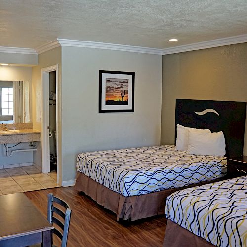 A hotel room with two beds, a desk, a TV, and a bathroom area. There's a painting on the wall and bedside lamps.