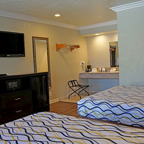 The image shows a hotel room with two beds, a TV on the wall, a small table with chairs, a dresser, a mirror, and a sink area with amenities.